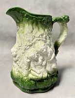 Front angled view showing bas-relief cherub scene with green and white glaze, ornate handle and scalloped base.