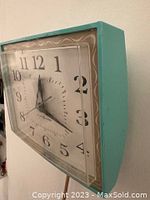 Front angle view showing the turquoise plastic frame and black numbered dial with three clock hands.