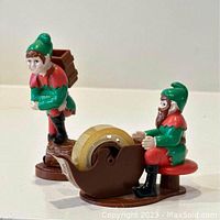 Frontal photo showing both elf figures: sitting elf tape dispenser with tape roll and standing elf with back satchel