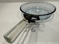 Top-down and angled view of vintage 1960's Pyrex glass frying pan, showing clear blue-tinted glass body with stainless steel trim and attached thick glass handle secured by a dark metal bracket.