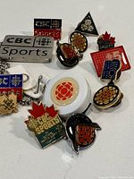 Collection of vintage CBC Sports lapel pins and keychain items laid out on a white background showing various pins with CBC logos and sports themes, as well as a clear keychain and a white key fob.