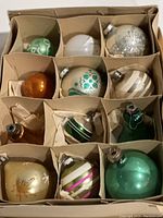 Overview of 12 vintage Christmas ornaments in original box with divided compartments showing various colors and designs including stripes, glitter floral and bell shapes.
