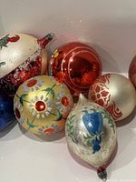 Group photo showing assortment of hand-painted vintage glass ornaments including yellow floral sphere, red shiny sphere with pine design, blue and white pear-shaped ornaments with floral designs