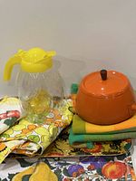 Overall view of the kitchen lot showing the glass jug with yellow lid, orange pot on folded cloth, floral textiles