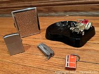 Photo showing vintage black glass Lensey ashtray with miniature classic car, Zippo lighter, textured smoking case and lighter tool on a white surface.