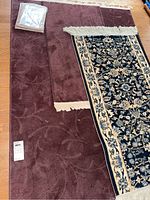 Photo showing the two larger area rugs: a large solid burgundy rug with floral pattern and fringe, partially covered by a black and beige floral patterned runner with fringe.