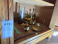 Overview photo of various brassware items inside a wooden cabinet shelf, includes candle holder, candlesticks, bell, keys, and other brass decorations.