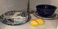 Ceramic plates under a lamp including a large blue floral patterned plate, smaller plates with the blue floral pattern, a dark blue ceramic bowl on a plate, and two yellow decorative lemons.