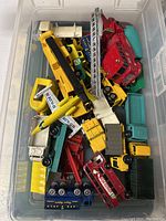 Top view of clear plastic bin containing mixed diecast vehicles including red fire truck, yellow crane truck, green BP truck and others