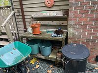 Wide shot showing resin and wood green wheelbarrow, black garbage can, planter pots under wooden bench, watering can on bench, boot cleaner on bench.