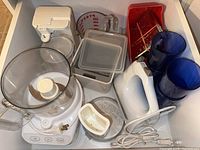 Top view of kitchen appliances and accessories including food processor, hand mixer, measuring cup, storage containers, utensil tray, and glasses.