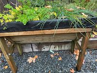 Full view of the wooden raised planter box with green plants growing inside, set on outdoor gravel.