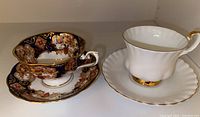 Two Royal Albert bone china teacups and saucers shown side by side on a shelf, one floral patterned with dark background and gold accents, the other white with gold trim.