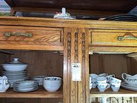Wooden cabinet shelf showing stacks of china including side plates, dinner plates, cups, bowls, and serving pieces with leaf and berry pattern.