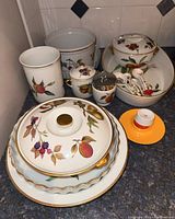 Overview of various Royal Worcester Evesham serveware arranged together showing plates, serving dishes, condiment jars, and bowls with spoons, including an orange candle holder.