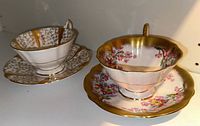 Two Queen Anne bone china teacups and saucers, one with floral pink and yellow pattern with gold trim and one with gold and leaf pattern.