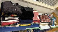 Overview of folded women's clothing stacked on table, includes sweaters, tops, and a plaid bag to the side