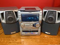 Front view of Aiwa CX-NAJ230 stereo with two speakers. Visible power button, volume knobs, display screen, and cassette decks.