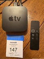 Top view of Apple TV device and full Apple TV remote on wooden surface with auction label.