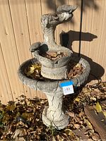 Full view of the concrete bird bath showing the entire structure from base to top, with leaves inside the bowls and a wooden fence backdrop.