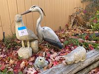 Four yard statues placed on ground covered with fall leaves: concrete seagull perched on bases, concrete frog sitting, concrete cat lying down, and tall plastic crane standing.