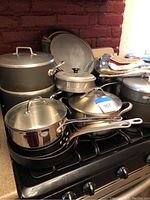 Overview image showing multiple pots and pans mostly with lids, stacked on a stove, with hot pads folded nearby and a large round pan leaning at the back.