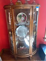 Full view of wooden curved glass display cabinet with three glass shelves containing decorative items (not included). Shows wood grain, curved glass sides, and front door.