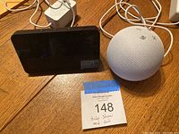 Photo shows a black Amazon Echo Show with an approximately 6-inch screen next to a white spherical Amazon Echo Dot smart speaker on a wooden table, along with power cords and a label indicating 'Echo Show and Jot'.