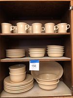 Photo of ivory Fiesta mugs on top shelf, stacked bowls and plates on middle and bottom shelves inside wooden cabinet.