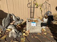 Photo showing kissing birds statue, concrete planter, water pump and cat statue on wooden deck with leaves.