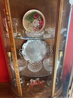 Glass and crystal items arranged on two shelves inside wooden cabinet showing serving plates and bowls, decorative plate, and glassware.