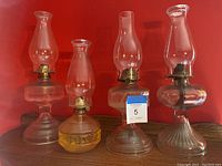 Photo showing four vintage glass oil lamps against a red background, with various base styles and clear chimneys.