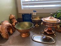 Photo of mixed metal housewares on countertop including copper colander, animal-shaped copper cake pans, brass pedestal bowl, copper chafing dish with glass insert, and small goblets in blue box.