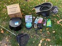 Wide view of all pond supplies, showing net, skimmer box, liquids, and plastic components on grass.