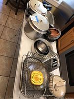 Overview image showing two large aluminum pressure cookers, nested metal pots and pans, wooden bowl, cheese grater, and wire racks on kitchen table.