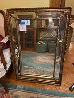 Largest gold-colored framed rectangular mirror with ornate details leaning against wall near upholstered chair.