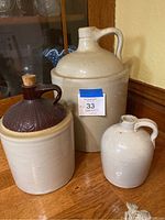 Three stoneware jugs displayed together on wood surface; includes large beige jug with handle, medium two-tone jug with cork, and small beige jug