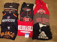 Nine folded men's sweatshirts piled showing team logos of Broncos, Nebraska, Cincinnati, Ohio State, and Adidas.