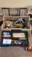 Photo showing open plastic toolbox with hand tools including caulking gun, saw, brushes, and other items.