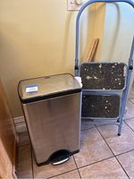 Stainless steel trash can and blue metal framed two-step stepstool side by side on tiled floor near wall