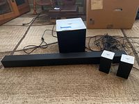 Sound bar system components arranged on carpeted floor: sound bar, subwoofer, two rear speakers, visible wires.