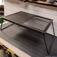 Side angled view of the campfire grill showing the foldable legs and combination of solid flat and mesh grilling surfaces.