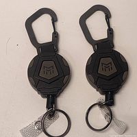 Two black retractable keychains with carabiner clips and keyrings laid flat on a white surface, top view showing their design and logos.