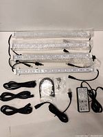 Four white metal LED light strips laid out with black power cords, power supply adapter, mounting hardware and connectors on white background.