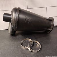 Black plastic cyclone dust collector separator positioned on its side with visible inlet and outlet connections. Two metal hose clamps placed in foreground.