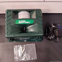 Electronic ultrasonic animal control device with motion sensor, green color, shown with power adapter and mounting screws laid out on a dark surface.