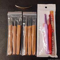 Photo showing five wooden-handled needlepoint or felting hooks, two plastic crochet hooks in packaging, and one small metal curved tool on a black background.