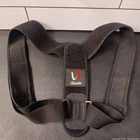 Top-down photo of a black posture corrector brace with padded wide straps and a central connecting piece featuring the Shiara logo. Adjustable Velcro strap visible.