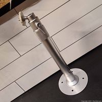 Silver metallic projector or camera mount bracket standing vertically showing round base plate with mounting holes and knurled adjustable section near the swivel head.
