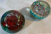 Two vintage glass paperweights side by side. Left is a multicolored swirl design paperweight with signature, right is a Millefiori floral patterned paper press ball.